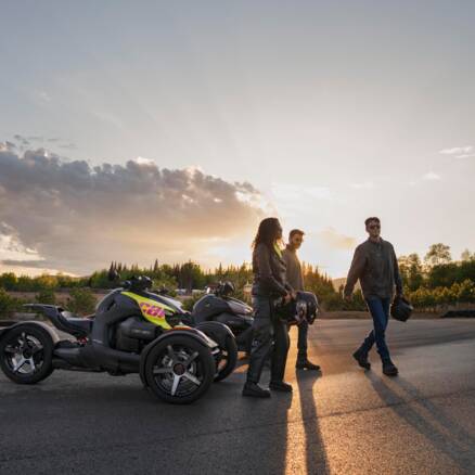 Zwei parkende Can-Am Ryker, drei Personen laufen vor den parkenden Fahrzeugen, im Hintergrund geht die Sonne unter
