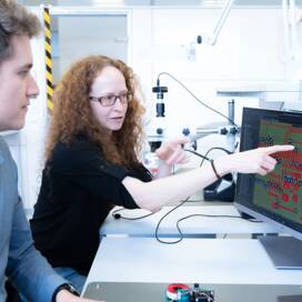Zwei Mitarbeiter:innen sitzen vor einem Computerbildschirm, Mitarbeiterin zeigt mit dem Finger auf den Bildschirm