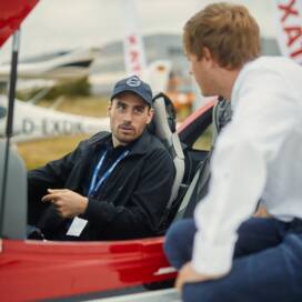 Zwei Männer unterhalten sich, einer sitzt in einem Flugzeug, der andere sitzt daneben, im Hintergrund sind Rotax-Flaggen zu sehen und weitere FLugzeuge