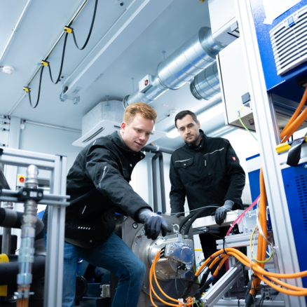 Eine Person in kompletter Werkstattbekleidung arbeitet an einem orangen HV Kabel unter dem Fahrzeug auf der Hebebühne, Blick Richtung Kabel. Ein zweiter Kollege assistiert und übergibt ein Werkzeug